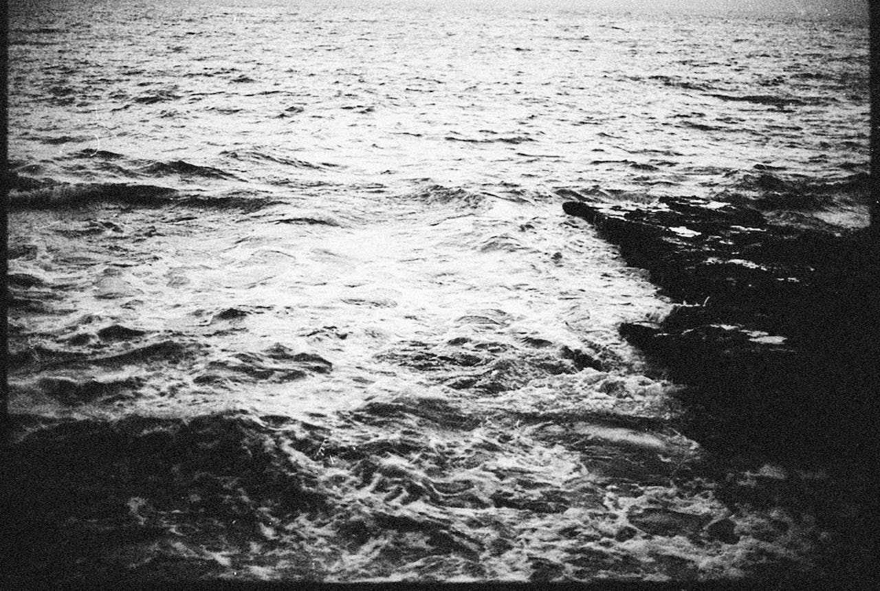 about-us-01 A moody black and white photograph captures waves crashing against rocky shore.