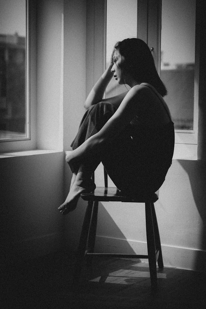 how-we-are A woman sitting on a chair by a window, deep in thought, creating a serene mood.