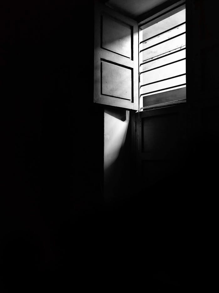 A black and white photo of an open window casting dramatic light and shadows indoors.