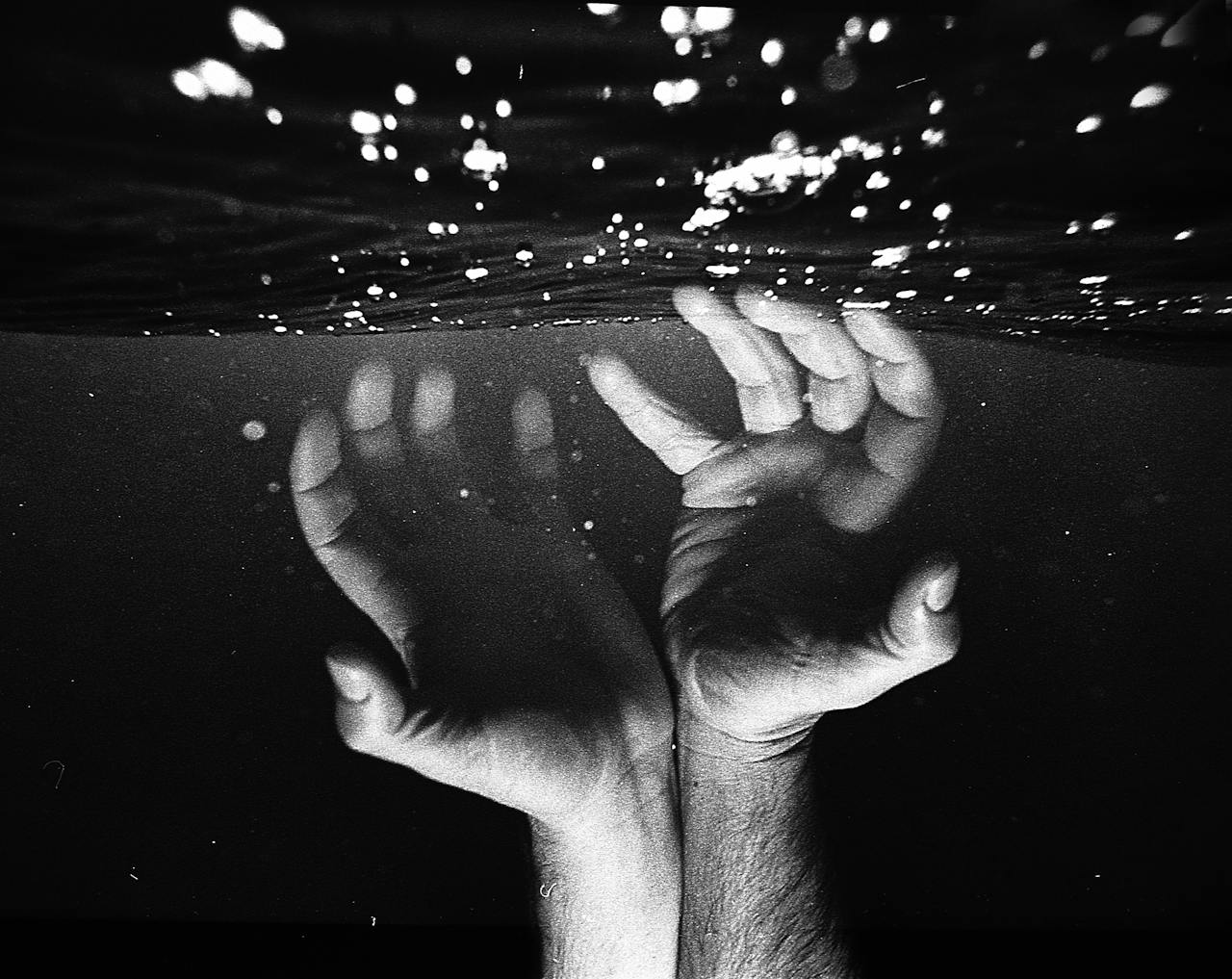 Black and white photo of hands reaching above the water with dramatic lighting.
